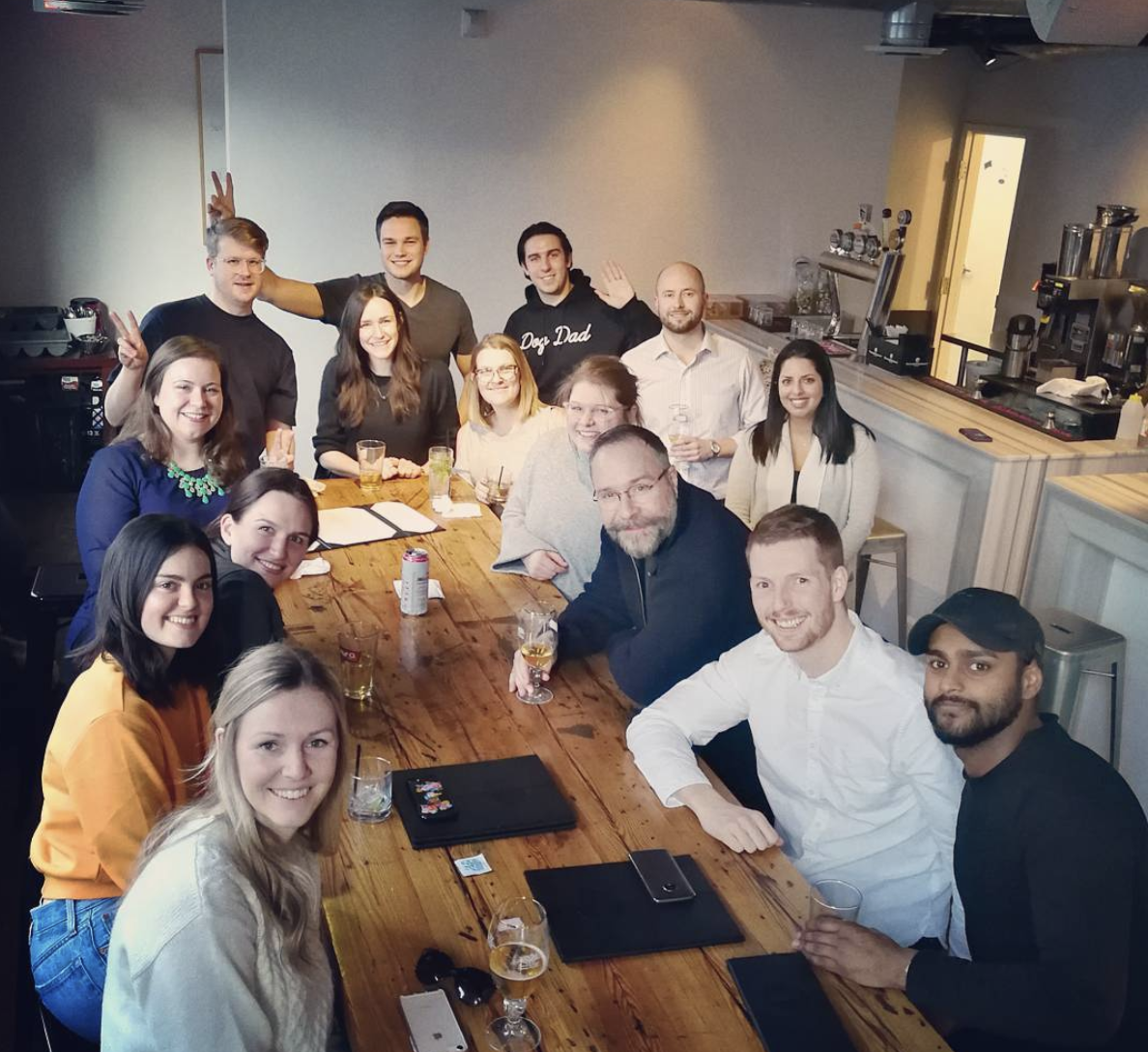 The PathFactory Team at a restaurant table with 15 people around the table all leaning in and smiling at the camera.